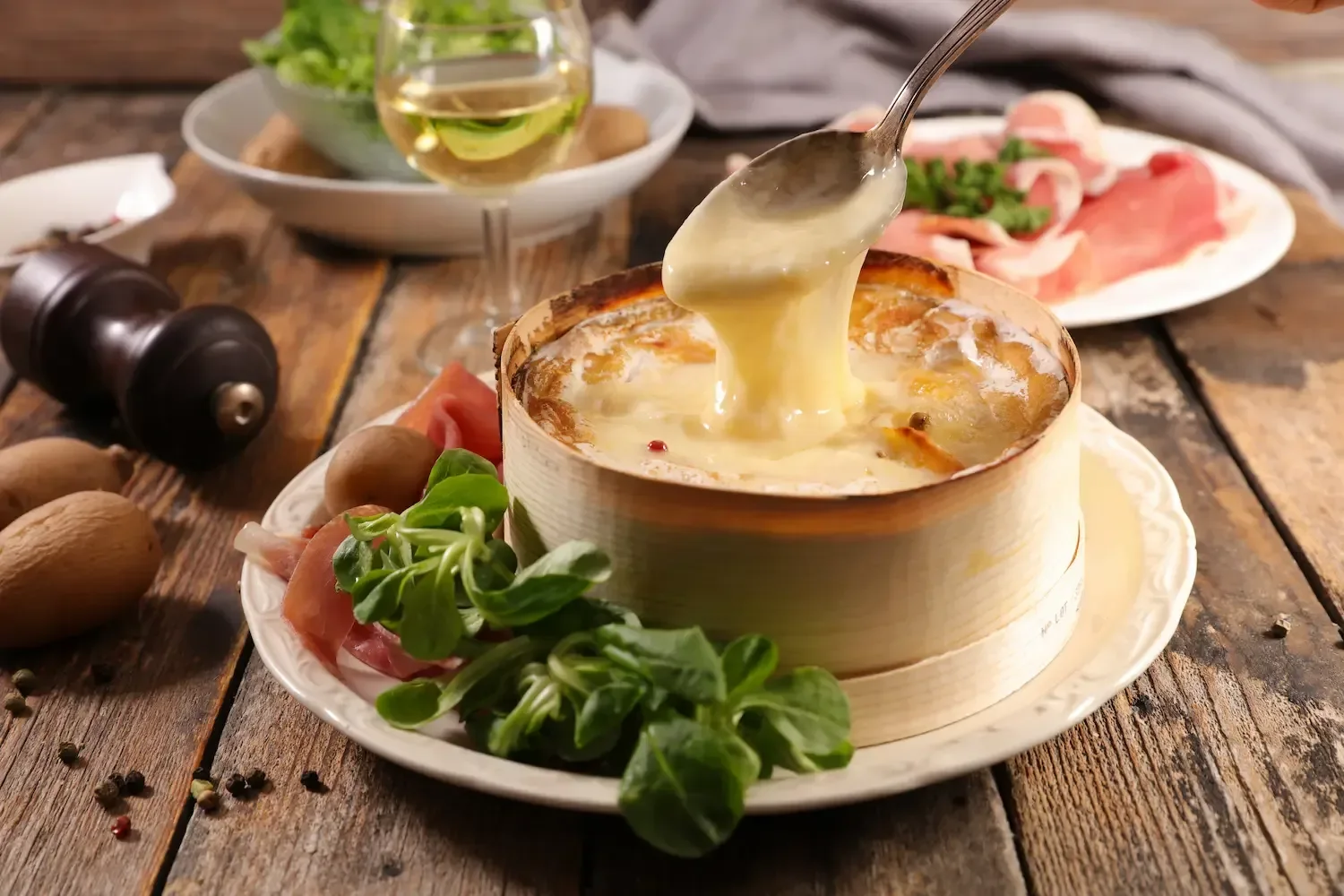 Un Mont d'Or sorti du four dans sa boîte en bois, fromage coulant levé à la cuillère, entouré de vin blanc, de charcuterie et de pommes de terre