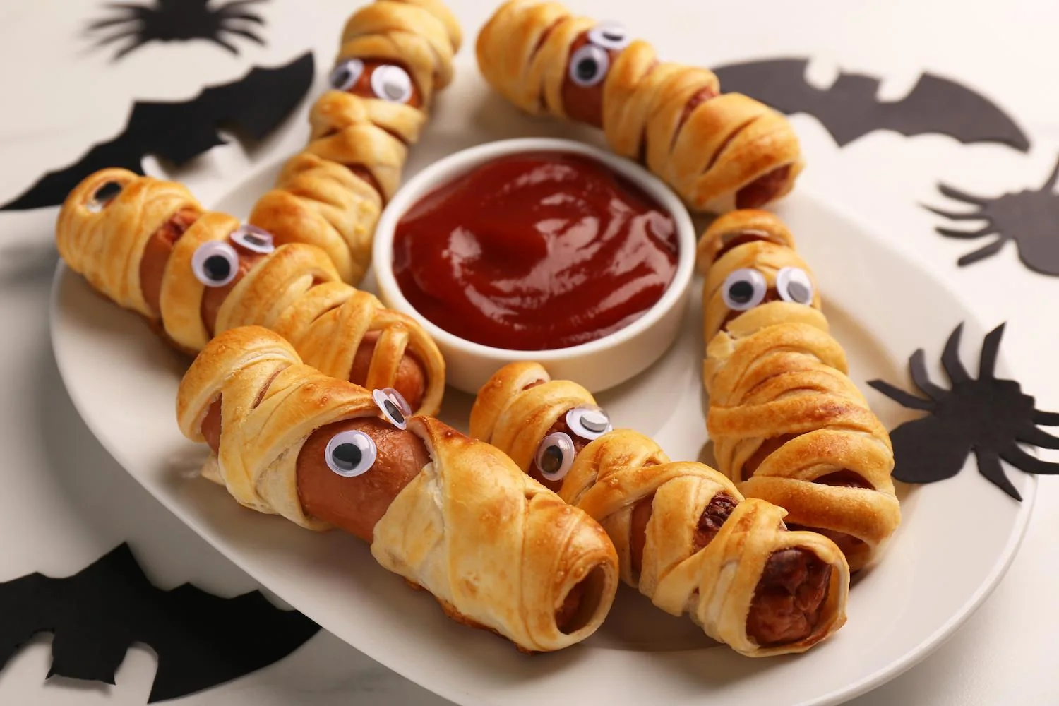 Des saucisses enroulées de bandelettes de pâte feuilletée, avec deux yeux en bonbon, servies sur une assiette blanche avec une sauce rouge